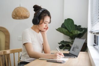 A woman sitting at her kitchen table, wearing headphones and working on a laptop.